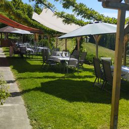 Ein sonniges Restaurant im Freien mit auf dem Rasen aufgestellten Tischen und Stühlen. Schirme und Markisen spenden Schatten. Die Gäste sitzen und genießen die Mahlzeit.