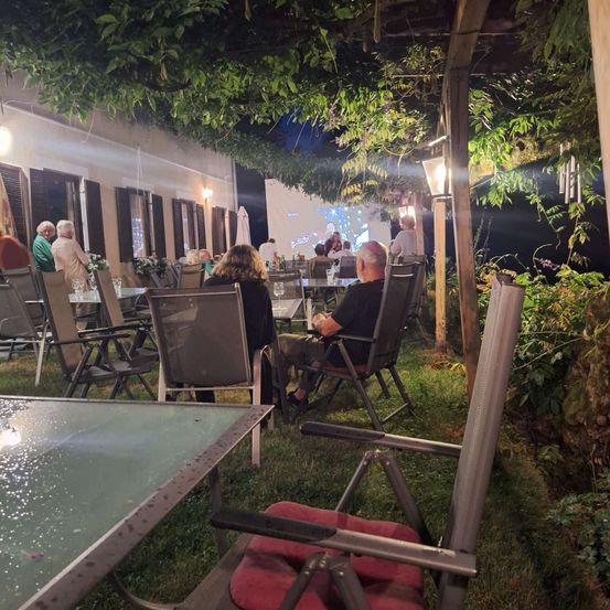 Eine Gruppe von Menschen sitzt im Freien in einem Restaurant unter einem Pergola mit einer Projektionsleinwand im Hintergrund.