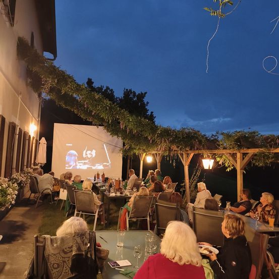Eine Gruppe von Menschen sitzt um Tische unter einem Pergola herum und schaut sich einen Film auf einer Projektionsleinwand an. Die Umgebung ist im Freien, mit Pflanzen und Bäumen im Hintergrund.