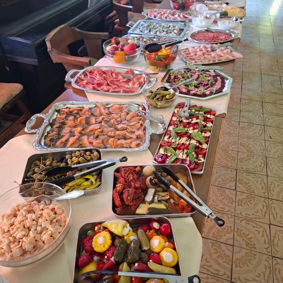 Ein langer Tisch mit verschiedenen Gerichten, darunter Fleisch, Gemüse und Obst. Es sind auch Schalen mit Garnelen und Oliven vorhanden.