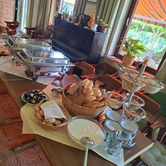 Ein Esstisch ist mit Brot, Gebäck und verschiedenen Gerichten gedeckt. Ein Buffettisch hält Chafing-Dishes und Schüsseln mit Essen. Ein Klavier und Topfpflanzen sind im Hintergrund.