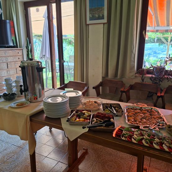 Ein hölzerner Tisch ist mit verschiedenen Gerichten gedeckt, darunter Teller, Schüsseln und Tabletts mit Essen. Eine Kaffeekanne und ein Kaktus stehen links, und Stühle sind um den Tisch herum angeordnet. Der Raum hat Glastüren, die zu einer Terrasse mit einem Tisch und Stühlen führen.