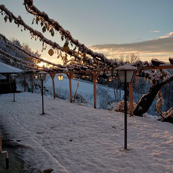 Eine verschneite Landschaft mit einem Gehweg, Holzstrukturen und Laternen. Die Sonne geht hinter einem verschneiten Hügel unter.