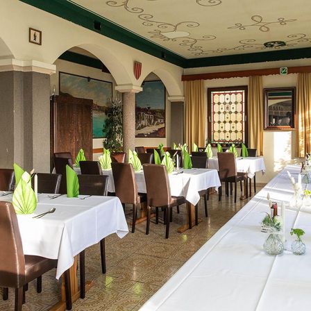 Ein Restaurant mit einem Essbereich, der aus Holzstühlen und Tischen mit weißen Tischdecken besteht. Der Raum hat einen gefliesten Boden und Wände, die mit Gemälden und einem dekorativen Fenster verziert sind.