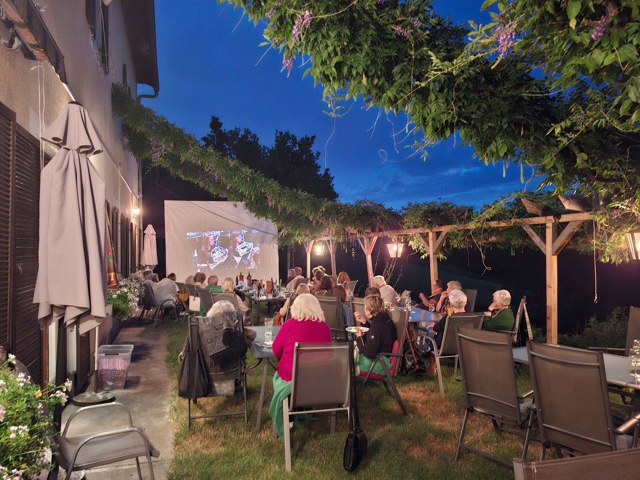 An outdoor movie screening at night with people seated under a pergola, watching a film on a projector screen.