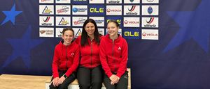 Drei Frauen in roten Jacken sitzen auf einer Bank vor einem blauen Hintergrund mit verschiedenen Logos. Der Hintergrund trägt die Aufschrift '2025 UCE Indoor Juniors European Championships Dad Hungary'.