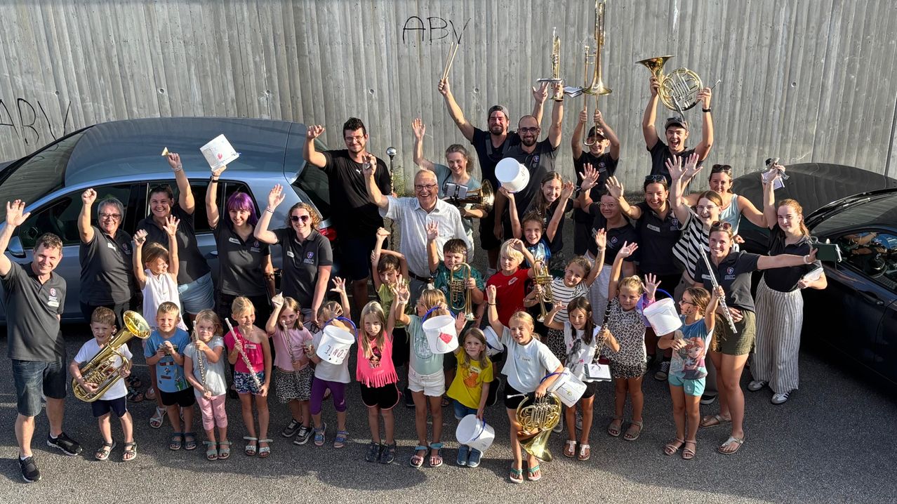 Eine Gruppe von Menschen, einschließlich Erwachsener und Kindern, versammelt sich draußen und posiert für ein Foto. Sie halten Musikinstrumente wie Trompeten und Tuben. Sie alle lächeln und winken. Dahinter befindet sich eine Wand mit Graffiti und ein geparktes Auto.