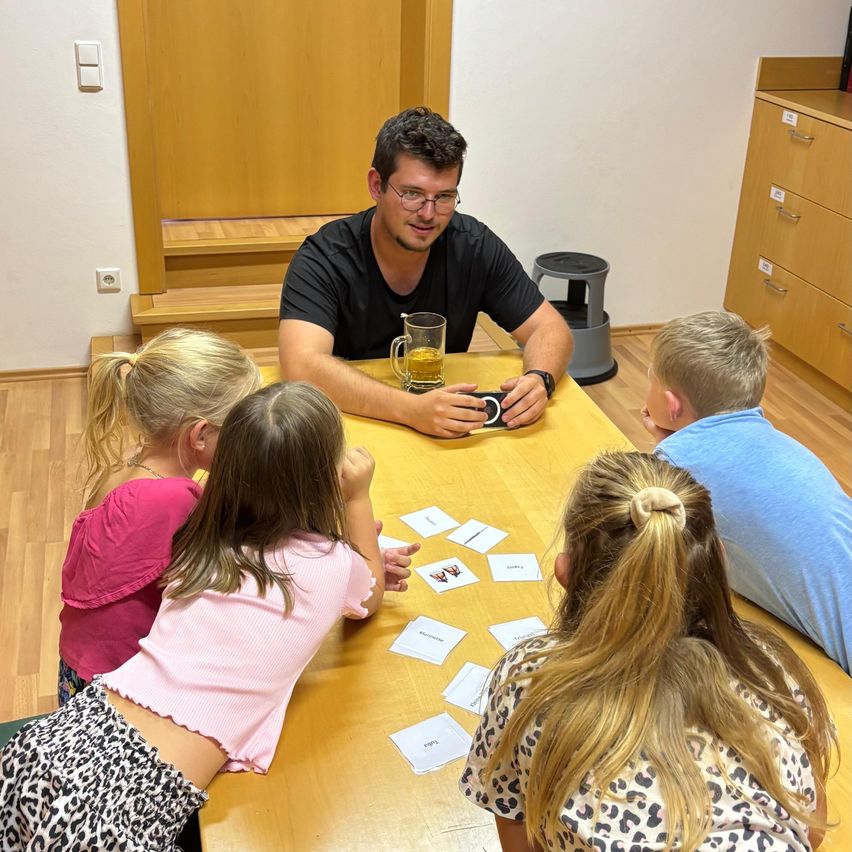 Ein Mann sitzt mit vier Kindern am Tisch, hält ein Telefon und ein Glas Getränk. Die Kinder spielen ein Kartenspiel mit Karten auf dem Tisch.