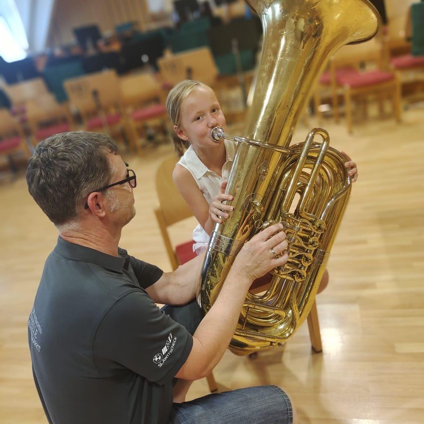 Ein Mann bringt einem kleinen Mädchen das Spielen einer Tuba in einem Raum mit Holzböden und Stühlen bei.