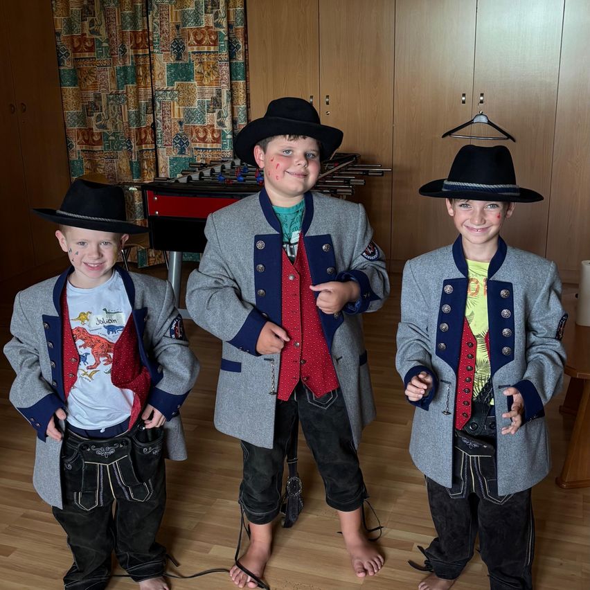 Drei junge Jungen in traditioneller bayerischer Kleidung mit roten Gesichtern, die in einem Raum mit Holzboden und Schränken im Hintergrund stehen.