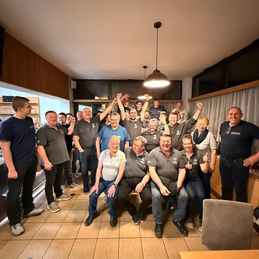 Eine Gruppe von Menschen in gleichen Shirts posiert für ein Foto, einige stehend und andere sitzend, in einem Raum mit Holzboden und Deckenlampen.