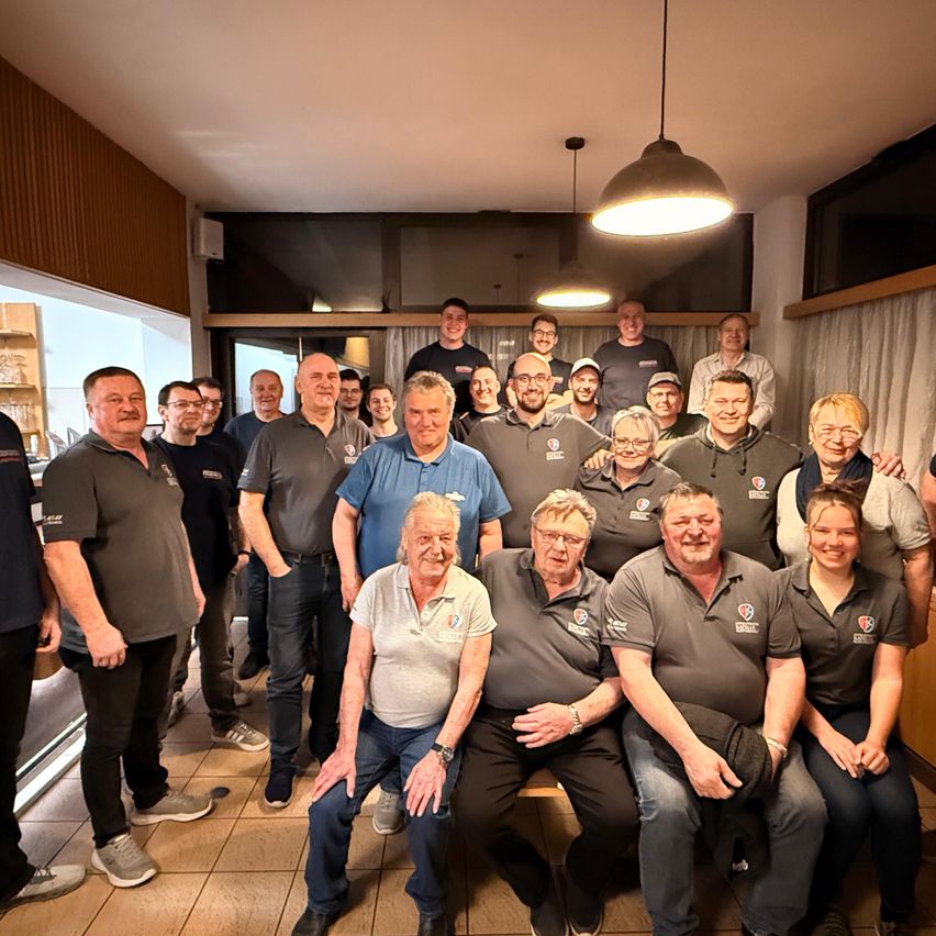 Eine Gruppe von Menschen, hauptsächlich Männer, stehen und sitzen in einem Raum, wahrscheinlich einem Restaurant. Sie tragen Uniformen mit Logos und einige tragen Brillen.