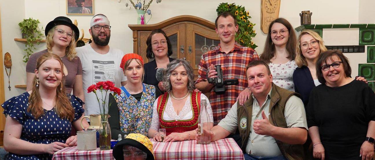 Eine Gruppe von Menschen in einem Raum, einige sitzend an einem Tisch mit einer Puppe, andere stehend, alle lächeln. Ein Mann mit einer Kamera steht hinter der sitzenden Gruppe.
