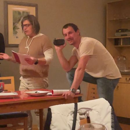 Ein Mann in einem weißen T-Shirt und Jeans macht ein Foto mit einer Kamera, während eine Frau mit Brille und Kette aus einem Blatt Papier liest.