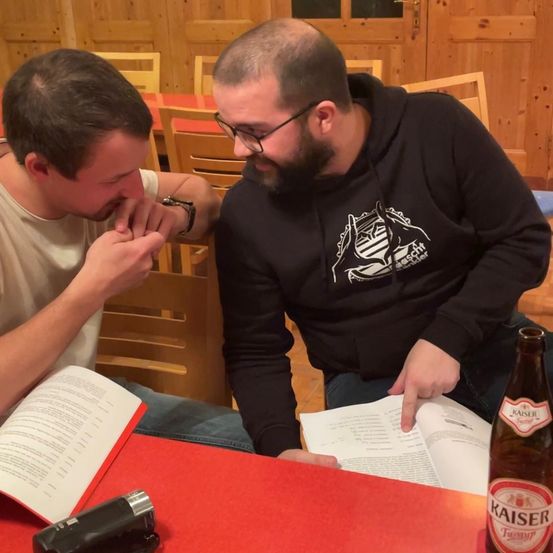 Zwei Männer sitzen an einem Tisch in einem Restaurant, einer liest ein Buch und der andere schaut hinein. Sie lächeln. Eine Flasche Kaiser-Bier steht auf dem Tisch.