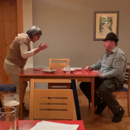 Eine Frau steht und gestikuliert mit ihren Händen, und ein Mann sitzt auf einem Stuhl, beide schauen sich an. Der Tisch hat zwei Gläser, eine Schüssel und einen Teller. Ein Bilderrahmen ist an der Wand montiert.