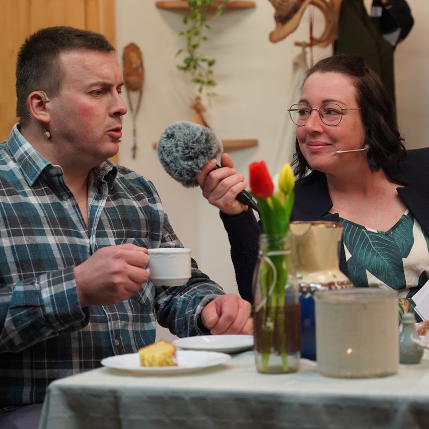 Ein Mann und eine Frau sitzen an einem Tisch mit Blumen und sprechen in ein Mikrofon. Der Mann hält eine Kaffeetasse, während die Frau lächelt. Hinter ihnen hängen verschiedene Gegenstände an der Wand.
