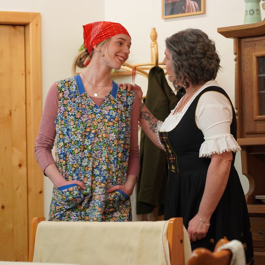 Zwei Frauen stehen in einem Raum. Eine trägt ein Blumenkleid und ein rotes Bandana, die andere ein schwarzes Kleid mit weißen Säumen. Hinter ihnen steht ein hölzerner Schrank mit einer Vase.