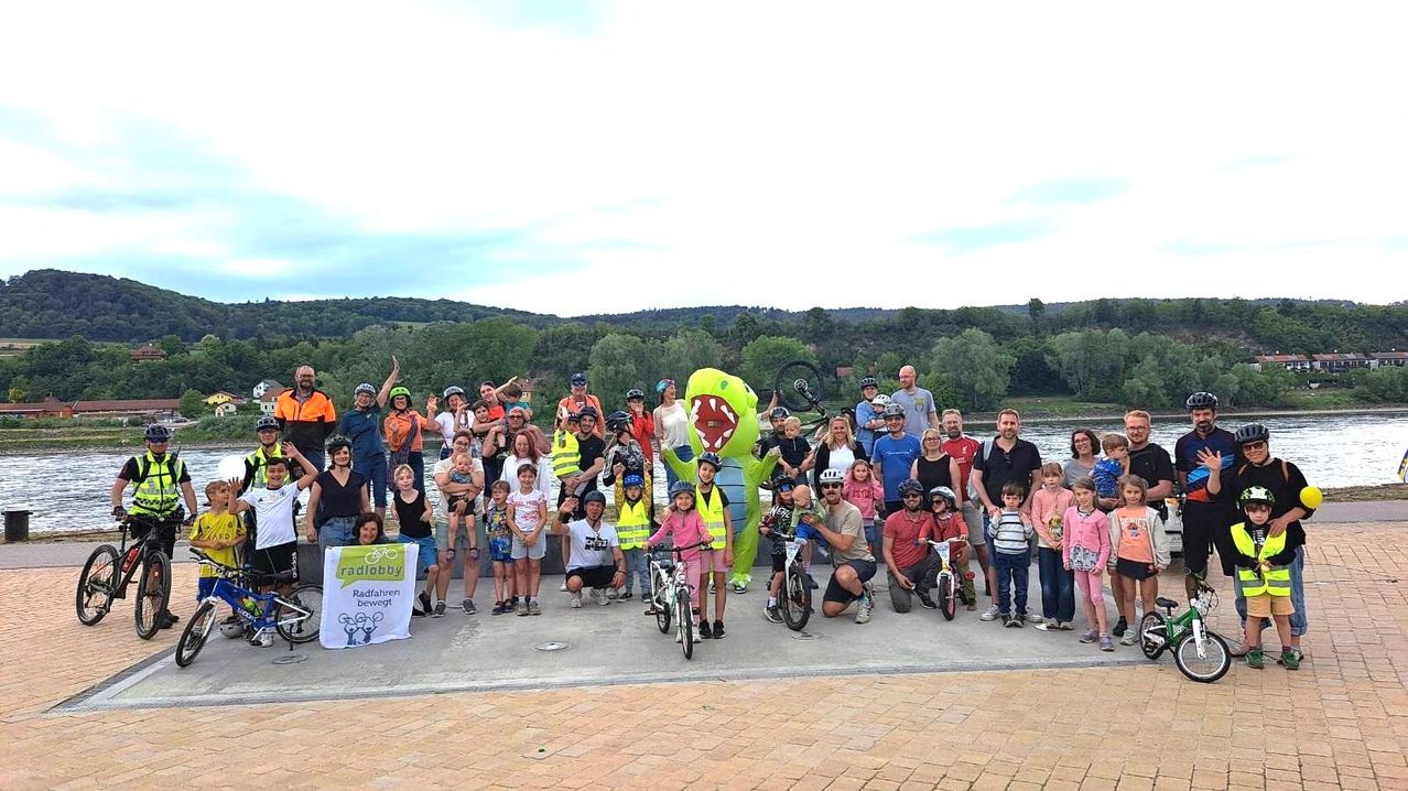 Eine große Gruppe von Menschen posiert für ein Foto im Freien mit Fahrrädern. Einige fahren und andere halten Kinder. Eine Maskottchen in der Mitte trägt ein Dinosaurierkostüm.