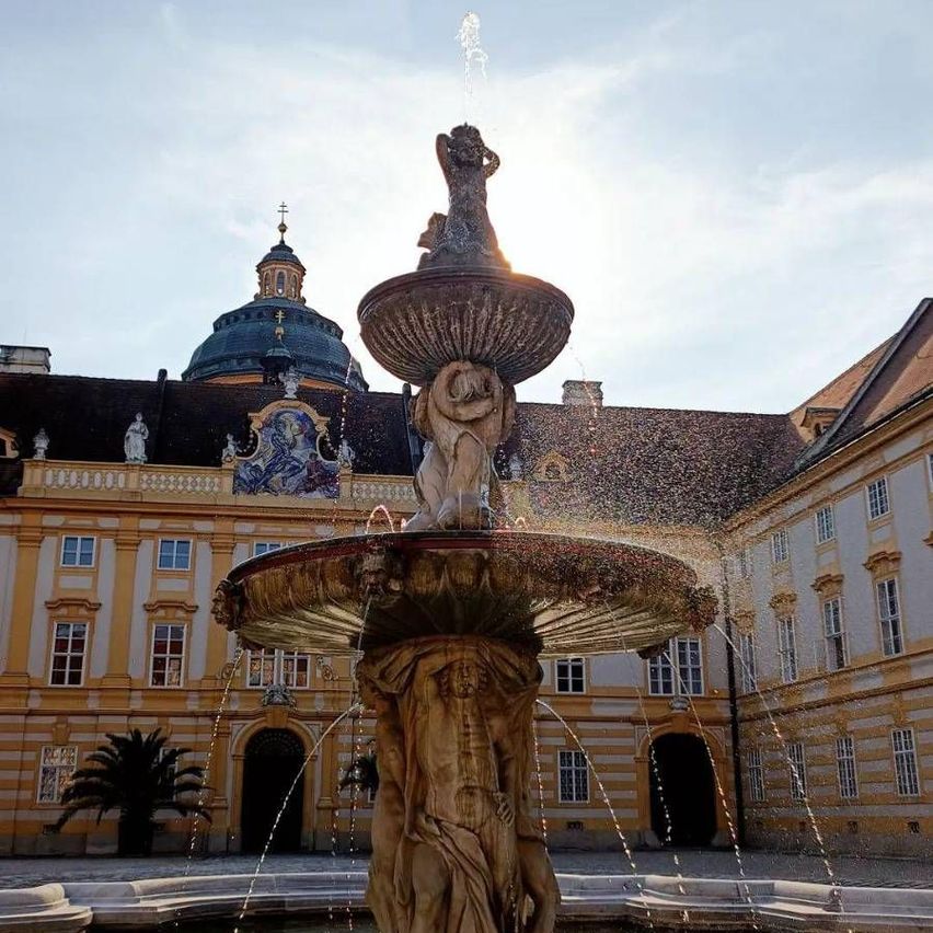 A large ornate fountain with statues stands in front of a grand building with a blue dome.