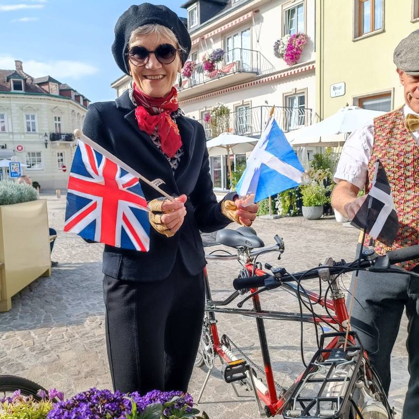 A woman in a black hat and sunglasses holds a British flag and smiles in front of a bicycle.
