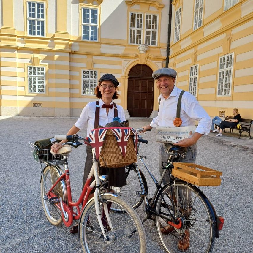 A man and a woman dressed in vintage clothing pose with their bicycles in front of a historic building.