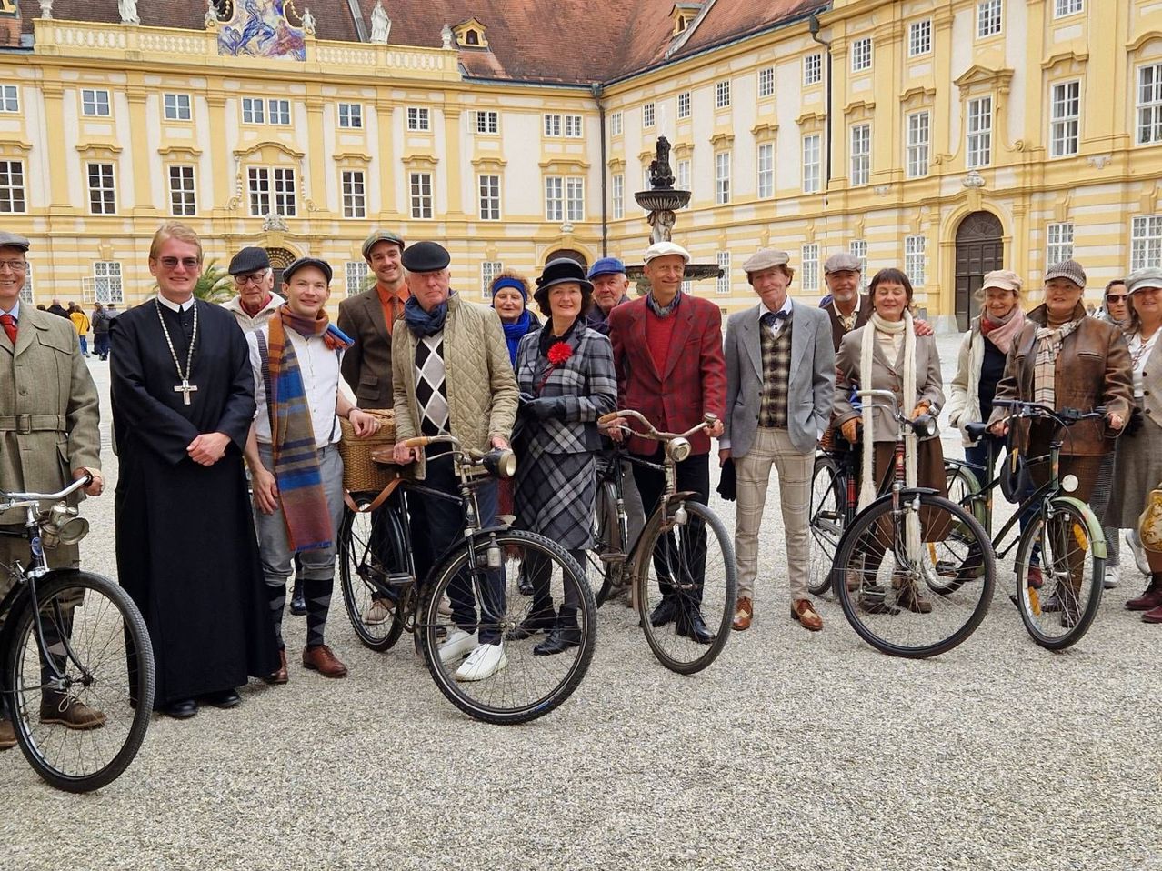 A group of people dressed in vintage clothing pose with their bicycles in front of a grand building.