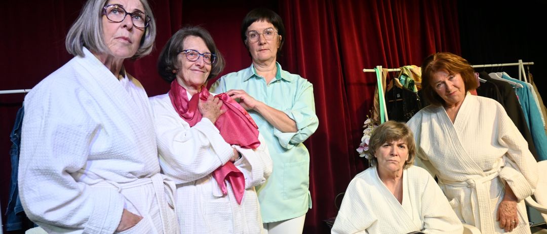 Fünf Frauen, in weißen Roben gekleidet, stehen gemeinsam vor einem roten Vorhang. Eine hält eine Zeitschrift, eine andere trägt einen rosa Schal, und die anderen haben verschiedene Posen eingenommen.