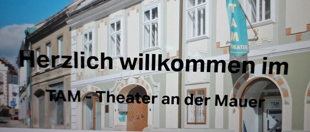 Ein Gebäude mit mehreren Fenstern und einer blauen Flagge. Der Text auf dem Gebäude lautet 'Herzlich willkommen TAM Theater an der Mauer'.