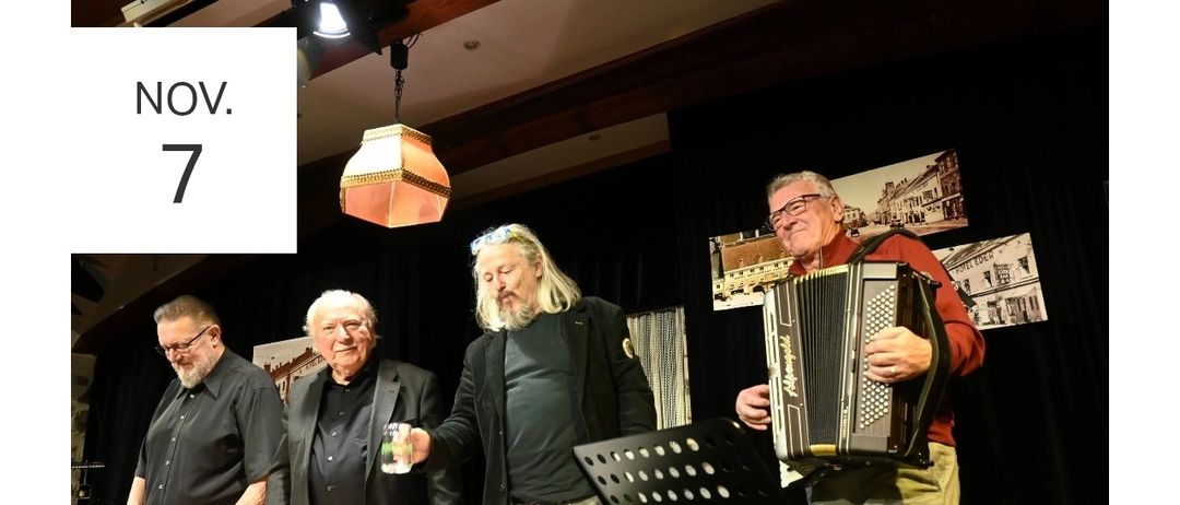 Vier Männer treten auf der Bühne auf. Der Mann in der Mitte hält ein Glas. Ein anderer Mann spielt Akkordeon. Ein Tisch mit grüner Tischdecke steht vor ihnen.