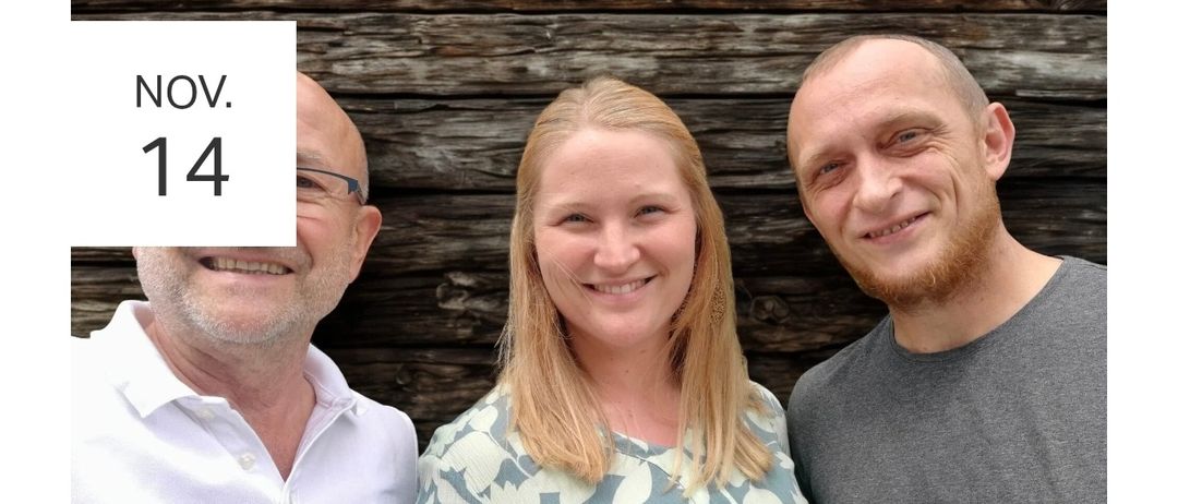 Drei Personen posieren für ein Foto vor einer Holzwand. Ein Mann links trägt eine Brille und ein weißes Poloshirt. Eine Frau in der Mitte lächelt mit einem Blusenkleid mit Blumenmuster. Ein bärtiger Mann rechts trägt ein graues T-Shirt.