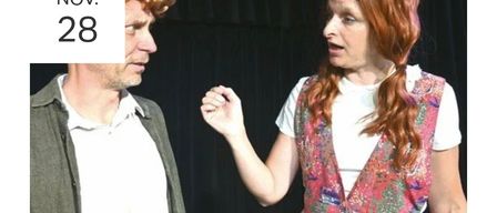 A man and a woman face each other, the woman is speaking and pointing her finger at the man, who is listening attentively. The woman has long curly red hair and is wearing a white sleeveless top with a floral pattern.