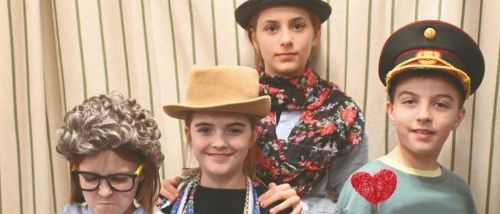 Fünf Kinder stehen zusammen in Kostümen. Das Kind auf der linken Seite hält eine Schachtel, die Mitte eine Schürze und der Junge auf der rechten Seite hält ein Herz und eine Zitrone.