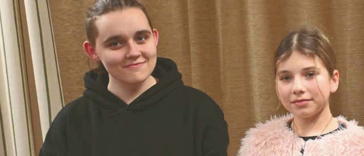 Zwei Mädchen, eines hält eine Babypuppe und das andere eine Geschenkbox, stehen gemeinsam vor einem Vorhang.