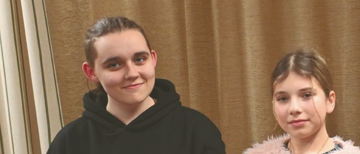 Zwei junge Mädchen posieren für ein Foto, eines hält eine in weißes Gewebe gewickelte Puppe und das andere eine Geschenkbox mit Blumendesign. Sie stehen nah beieinander vor einem Vorhang im Hintergrund.