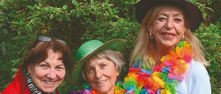 Drei Frauen stehen zusammen vor einem buschigen Hintergrund. Die Frau auf der linken Seite trägt eine rote Jacke und einen grünen Hut. Die Frau in der Mitte trägt einen grünen Hut und eine Girlande. Die Frau auf der rechten Seite trägt ein weißes Hemd und einen lila Hut mit einer Girlande.