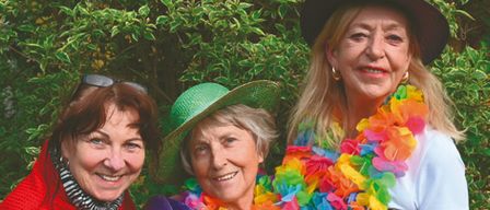 Drei Frauen stehen lächelnd vor einem Busch. Die Frau auf der linken Seite trägt einen roten Pullover, die Frau in der Mitte trägt einen grünen Hut und die Frau auf der rechten Seite trägt eine bunte Lei. Die Frau auf der rechten Seite trägt auch einen Hut und hält einen roten Hut.