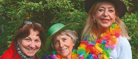 Drei Frauen stehen lächelnd vor einem Busch. Die Frau auf der linken Seite trägt einen roten Pullover, die Frau in der Mitte trägt einen grünen Hut und die Frau auf der rechten Seite trägt eine bunte Lei. Die Frau auf der rechten Seite trägt auch einen Hut und hält einen roten Hut.