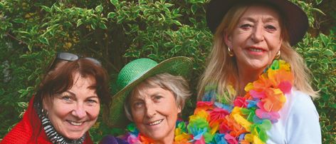 Drei Frauen in farbenfroher Kleidung, einschließlich Hüten und Blumenkränzen, lächeln und posieren für ein Foto im Freien vor einem Hintergrund aus grünem Laub.