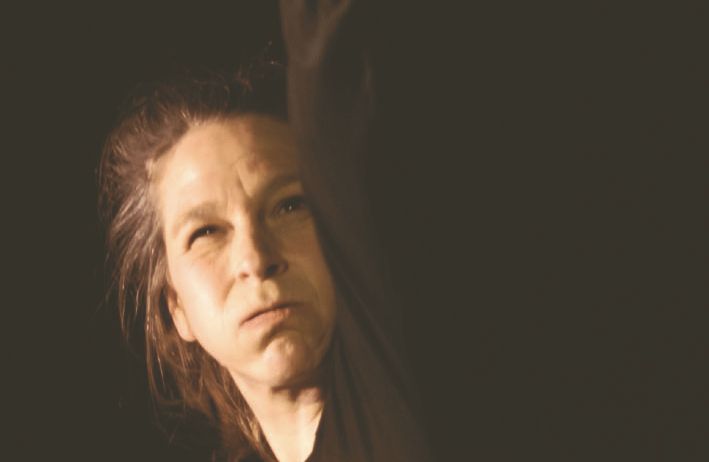 Eine Frau mit langen Haaren tritt auf der Bühne auf. Sie trägt ein schwarzes Langarmshirt und hat die Faust erhoben. Sie schaut mit ernster Miene nach unten. Der Hintergrund ist dunkel.