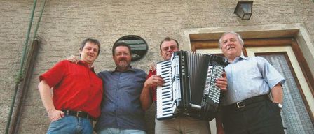 Four men stand in front of a building. The man on the right holds an accordion. Above them is a mural with a sign that reads 'Weinstube'.