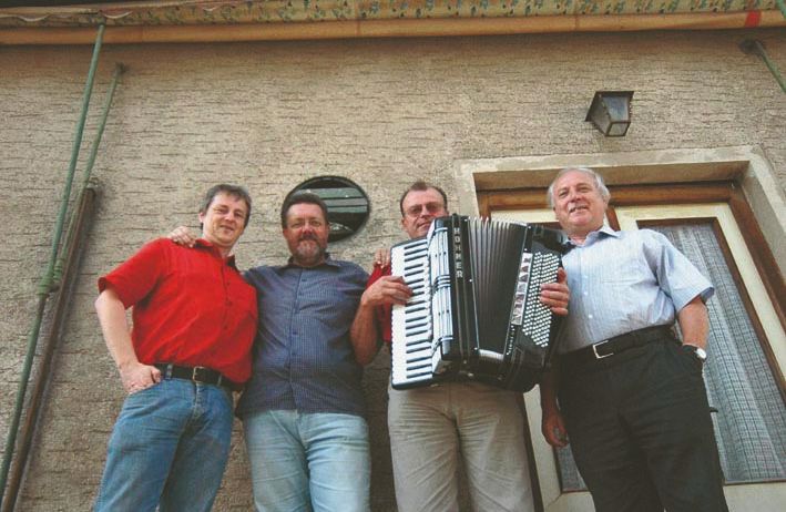 Vier Männer stehen vor einem Gebäude. Der Mann auf der rechten Seite hält ein Akkordeon. Über ihnen befindet sich eine Wandmalerei mit einem Schild, auf dem 'Weinstube' steht.