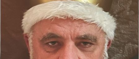 Ein Mann mit weißem Bart und Schnurrbart trägt eine goldene Krone, verziert mit Juwelen, und posiert ernsthaft vor einem braunen Hintergrund.