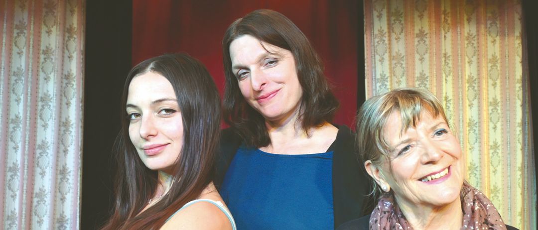 Drei Frauen lächeln und posieren für ein Foto. Eine in Blau, eine andere in Rot und die dritte trägt eine Schürze.