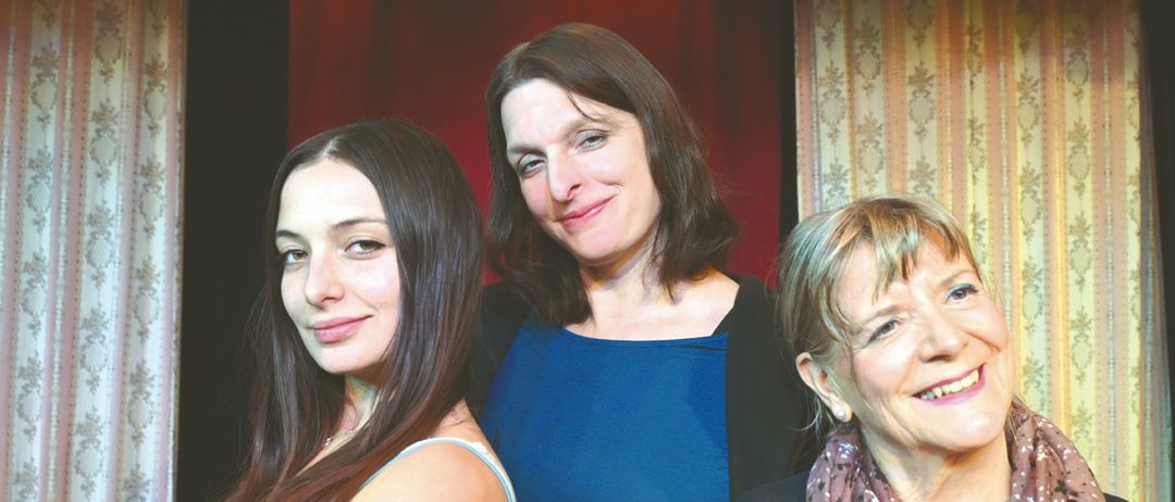 Drei Frauen posieren lächelnd zusammen. Die Frau auf der linken Seite trägt ein hellblaues Tanktop. Die Frau in der Mitte trägt ein dunkelblaues Oberteil. Die Frau auf der rechten Seite trägt ein kastanienbraunes Hemd mit einer Halskette.