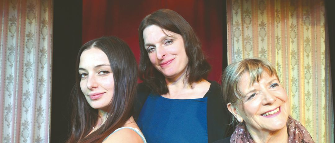 Drei Frauen posieren für ein Foto; eine in Blau, eine in Schwarz und eine in Rot, alle lächeln vor einem roten Vorhang.