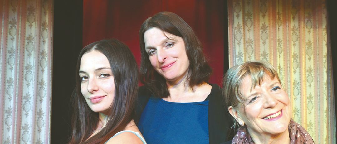 Drei Frauen stehen eng beieinander und lächeln für ein Foto vor einer gemusterten Wand. Die Frau auf der linken Seite trägt ein ärmelloses Top, die mittlere eine blaue Bluse und die rechte ein rotes Shirt und einen Schal.