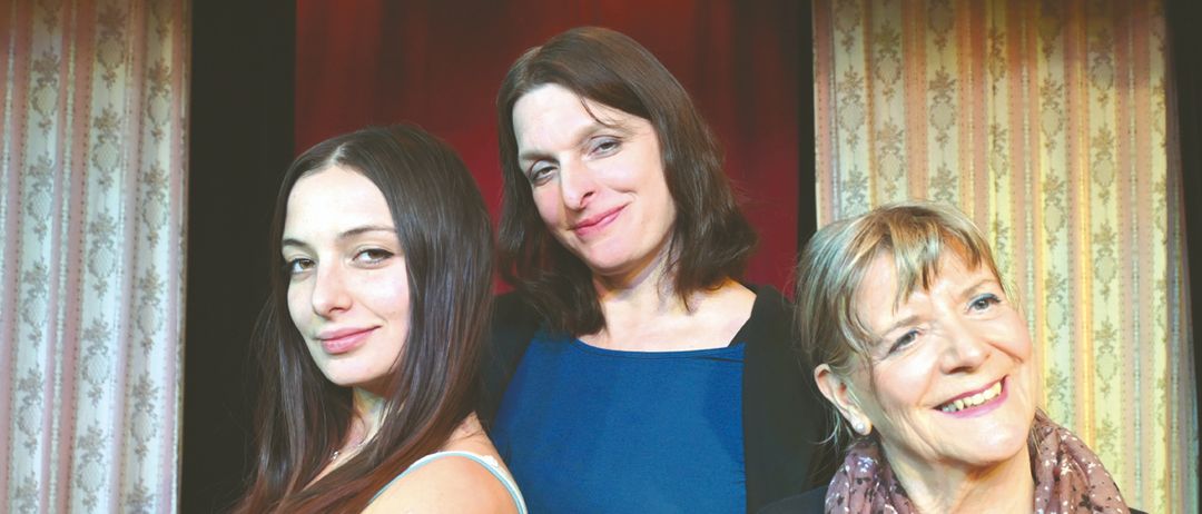 Drei Frauen lächeln für ein Foto. Die Frau auf der linken Seite trägt ein hellblaues ärmelloses Top. Die Frau in der Mitte trägt ein blaues Blusenshirt. Die Frau auf der rechten Seite trägt ein kastanienbraunes Oberteil und einen gemusterten Schal. Sie stehen vor einem roten Vorhang.