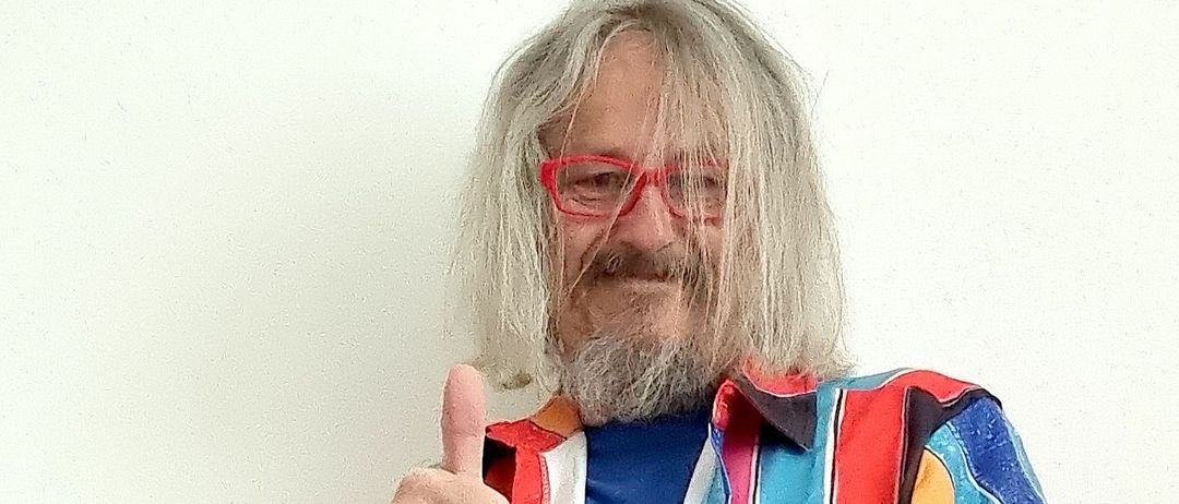 Ein Mann mit langen Haaren und Bart, der einen Daumen hochhält, trägt ein farbenfrohes Hemd und Jeans und steht vor einer weißen Wand.