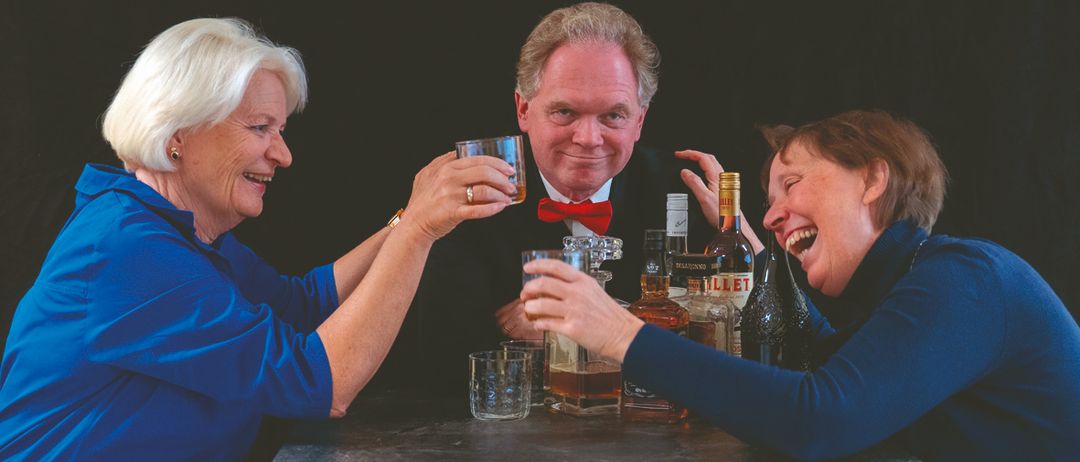 Drei Erwachsene in blauen Hemden versammeln sich um einen Tisch, lächeln und halten Gläser mit Alkohol in den Händen. Ein Mann in Anzug und rotem Fliege steht zwischen zwei Frauen. Flaschen mit Alkohol stehen auf dem Tisch.